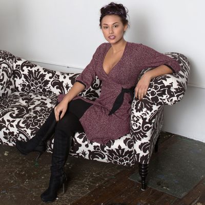 Petite woman sitting on patterned couch in wine diana wrap dress with black leggings